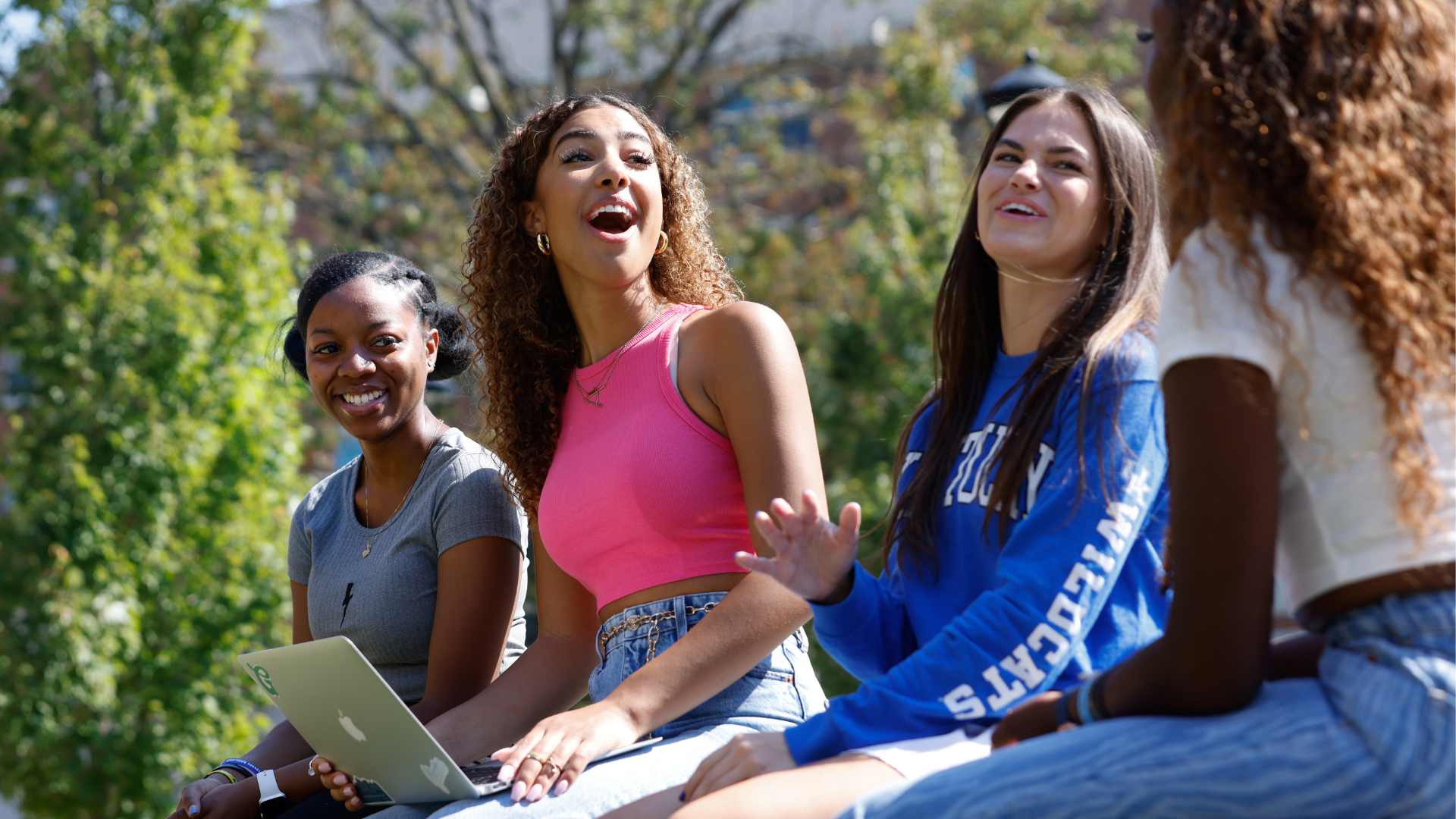 4 women sitting and laughing on campus bench, one holding a laptop
