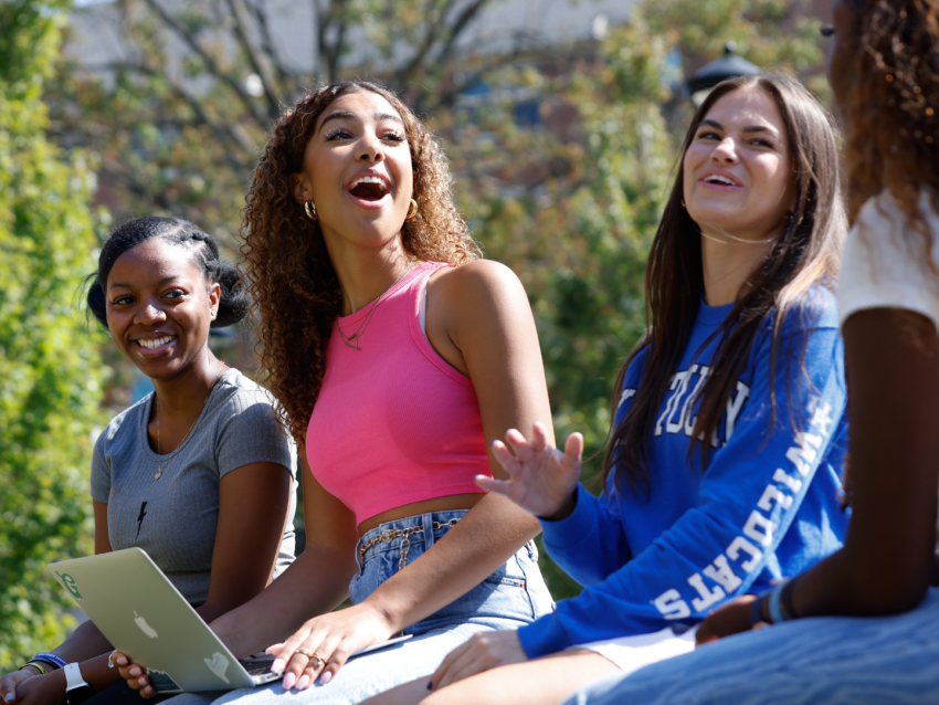 4 women sitting and laughing on campus bench, one holding a laptop
