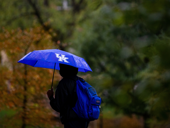 Student walking in front of trees with blue UK umbrella